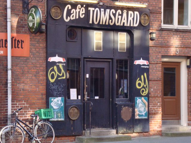 Her et nyere foto af cafe Tomsgården på Birkedommervej. Artikel fra Københavnerguiden:
Tomsgård er opkaldt efter den nærliggende Tomsgårdsvej som nok er lidt mere fashionabel og landskendt end Birkedommervej, men hvorfor i grunden det? Dette sted har egentlig ikke brug for reklame eller lånte fjer, for det har lige præcis et passende stampublikum som kan holde en stille og stabil omsætning. Det er også meget nemmere, for når alle ved hvad de kan forvente, er der jo ingen grund til at lefle for potentielle nye kunder med nymodens opfund som musik eller nogenlunde inspirerende udsmykning. I en menneskealder har her hængt en stor samling kasketter og en enkelt høj hat, og mere fis og ballade kan man virkelig ikke forvente. 